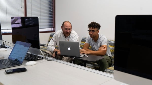 Two men, one college age and one older, sit and look at a laptop