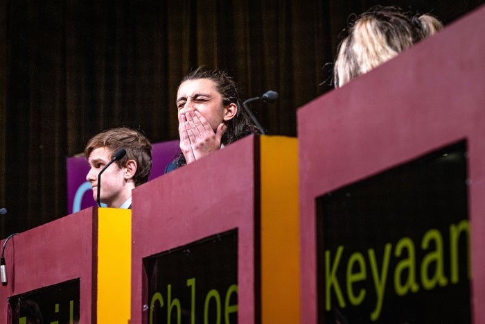 A college student behind a lectern scrunches up his face as he reacts to getting a question wrong