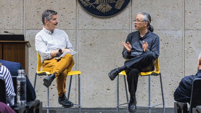 Two men on stools speak in front of an audience