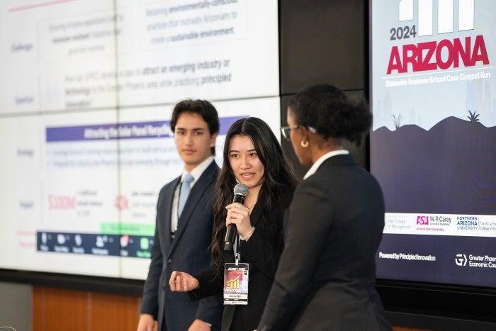 Three people standing in front of a large screen as one of them speaks into a microphone.