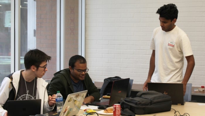 Two students sitting at a table and a third standing while they discuss computer science