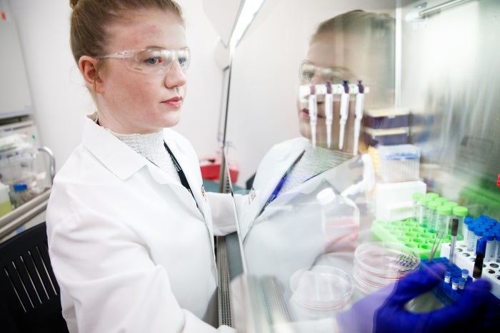 Colley wears a lab coat and goggles while handling lab equipment at a bench station.