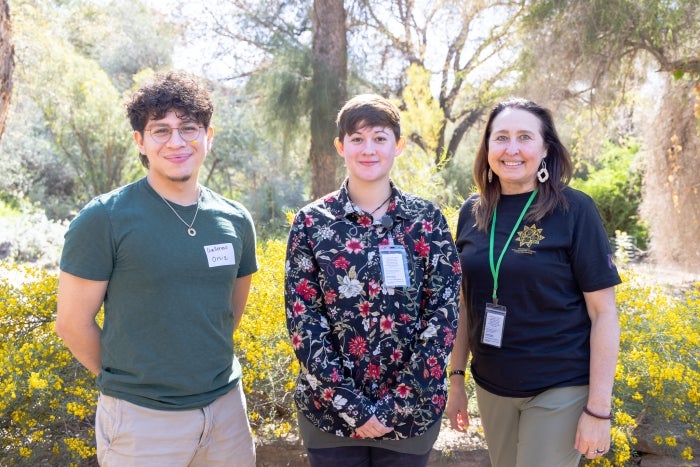 Three individuals smile at the camera in front of desert plants with yellow blooms.