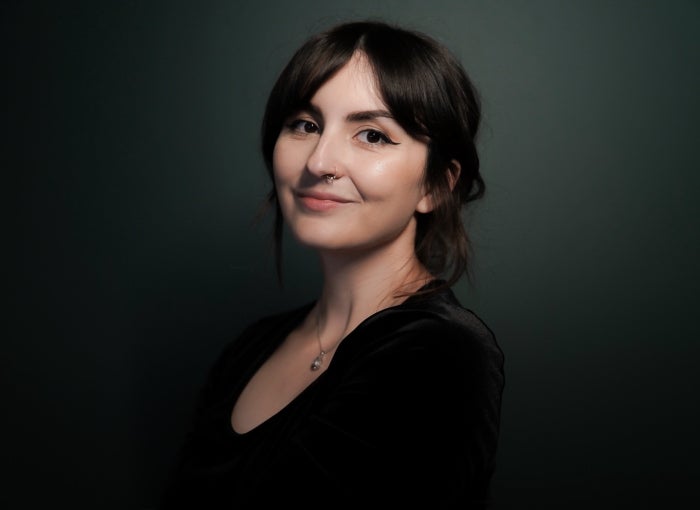 A woman stands against a dark green background while wearing a black top and a Mona Lisa smile.