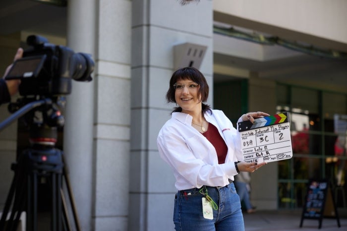 A student poses in front of a film camera with a clap board