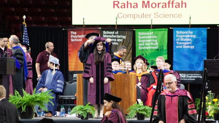 A student is hooded by a faculty member onstage at a graduation ceremony.