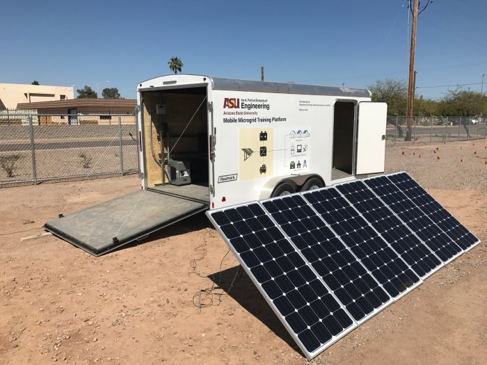 The mobile training trailer, a while trailer with back open, and a solar panel array in the desert landscape of the Polytechnic campus test bed