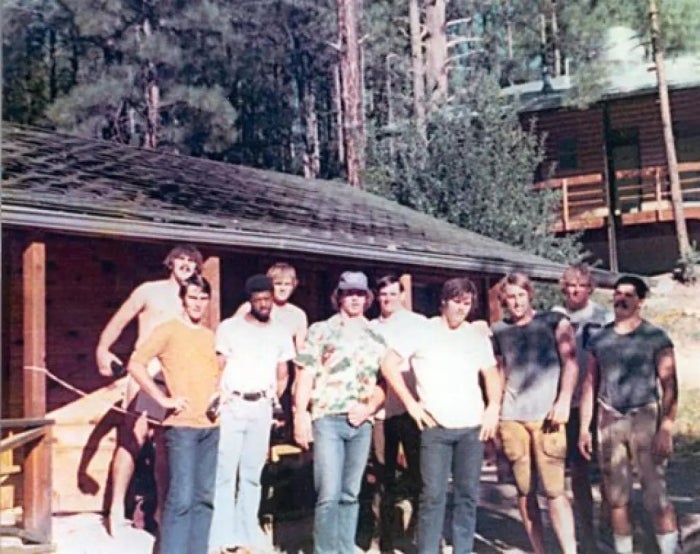 1970s photo of a group of men standing in front of a cabin in a pine forest