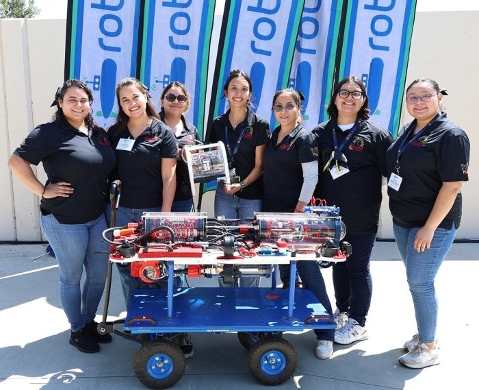 Group of seven women holding a small robot and standing behind a larger robot