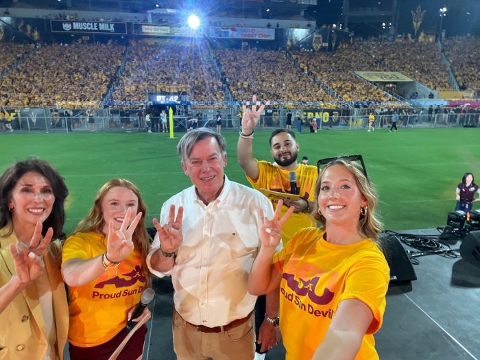 Five people pose for a selfie on the field of the ASU football stadium with a crowd of students in gold shirts behind them