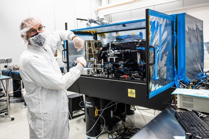 A man in clean room robes gestures toward a space instrument