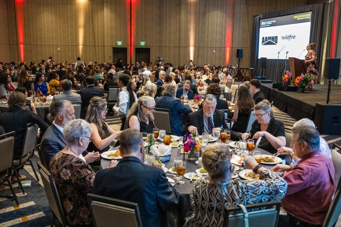 Audience seated at round tables listens to person on stage speaking