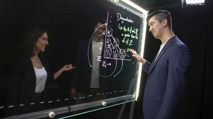 Faculty members work on a mathematical proof at a glass, lit board.