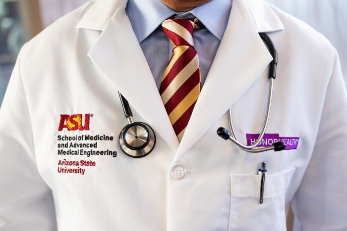 A man wears a doctor's lab coat with ASU and HonorHealth names embroidered on either side