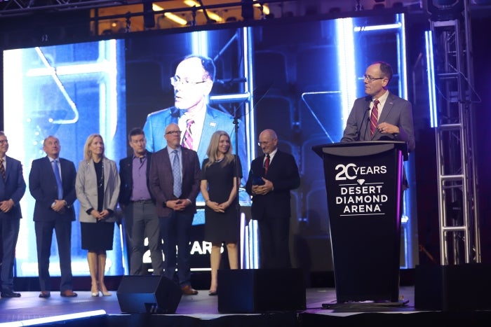 Man speaks at lectern at event while a line of people stand on stage behind him