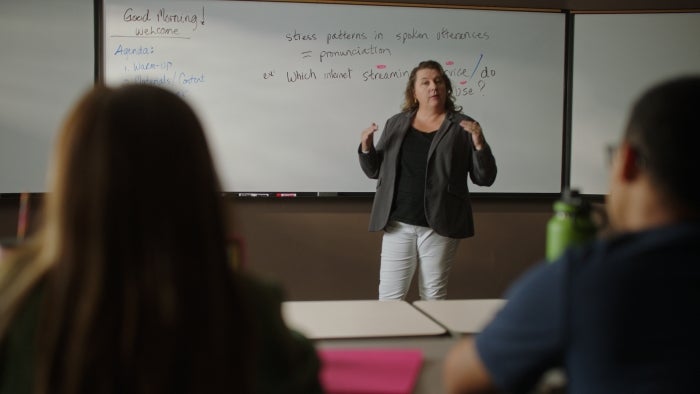 A teacher stands in front of a whiteboard, gesturing during a classroom lecture, with students in the foreground.