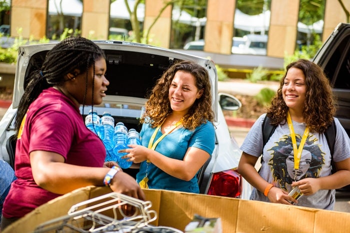 Three young women interacting by an open car trunk with water bottles outside a building during move-in week.