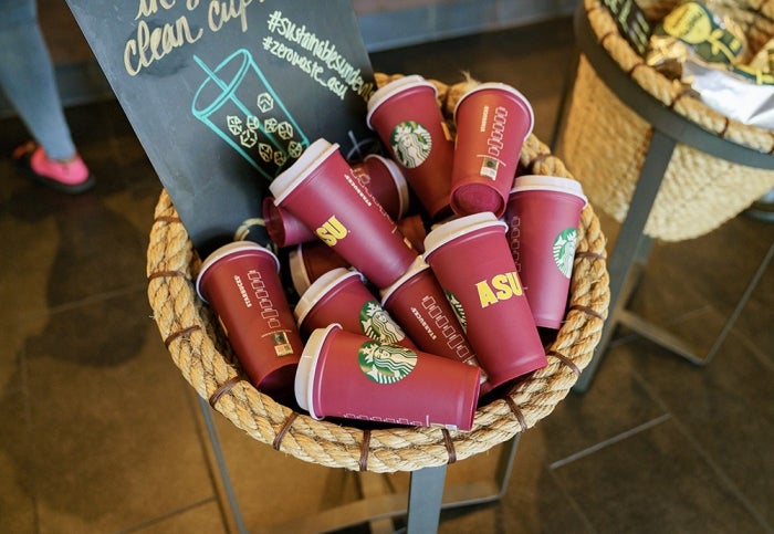 A basket filled with maroon reusable cups with Starbucks and ASU logos next to a blackboard illustration.