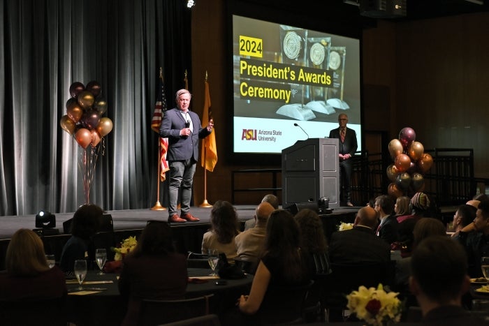 ASU President Crow stand on stage addressing crowd with projection sign behind him that says President's Awards Ceremony