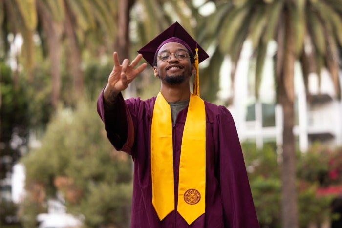 A young man in a maroon cap and graduation gown flashes the ASU pitchfork symbol with his right hand facing the camera