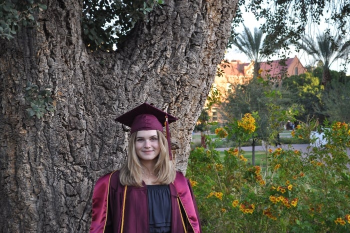 Young woman with medium length blonde hair wearing maroon graduation cap and gown poses in front of a tree