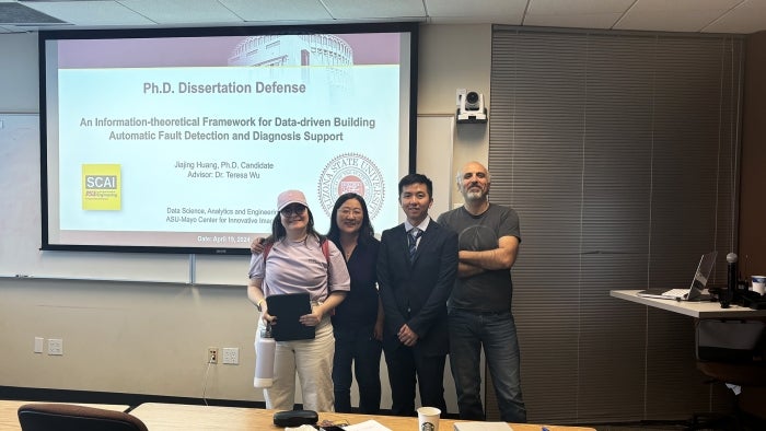 Jiajing Huang poses with faculty advisors in front of a screen.