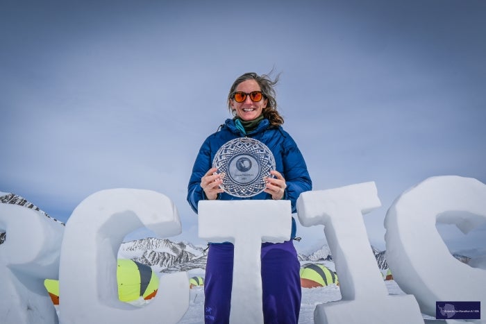 Julia Tizard Hunter holding a glass award in a snowy landscape with snow-covered letters spelling "Antarctica"