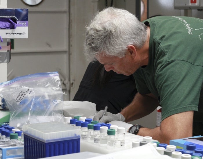 Man in a lab wearing gloves and taking notes next to several vials.