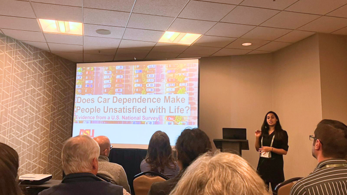 Woman pointing to a screen displaying a slide with information about car dependency and life satisfaction as she speaks to a room of people.