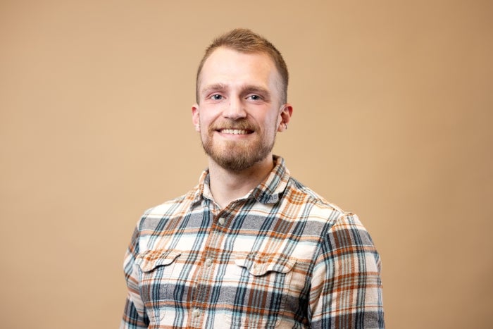 Portrait of a white man with short brown hair and a beard wearing a blue and brown plaid button-up shirt