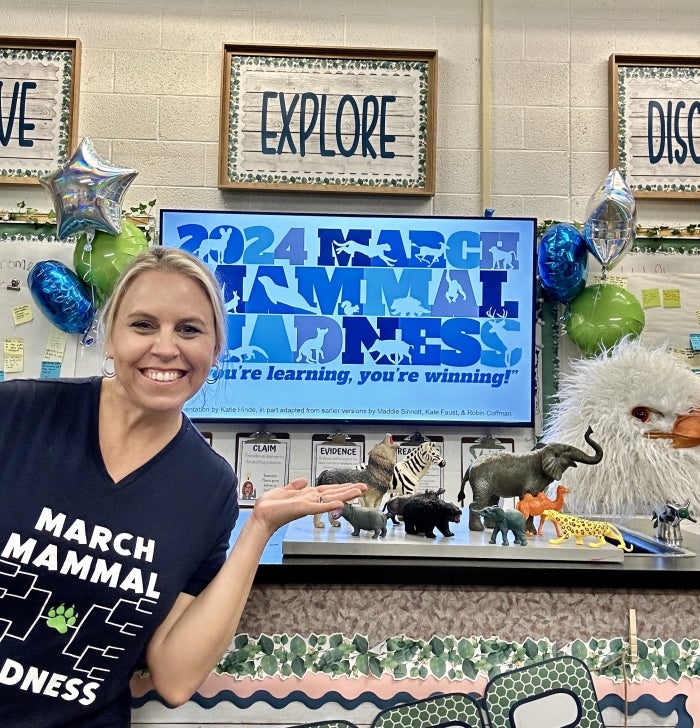 A woman in a t-shirt stands in a classroom with a "March Mammal Madness" screen and toy animals.