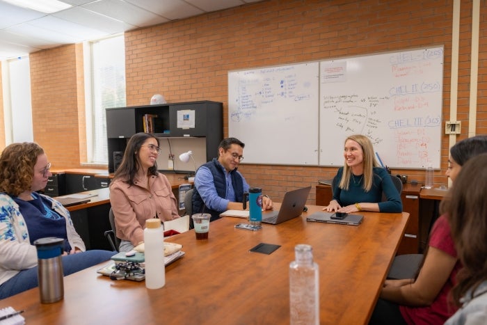 A group of people sitting around a table in a classroom with a white board behind them