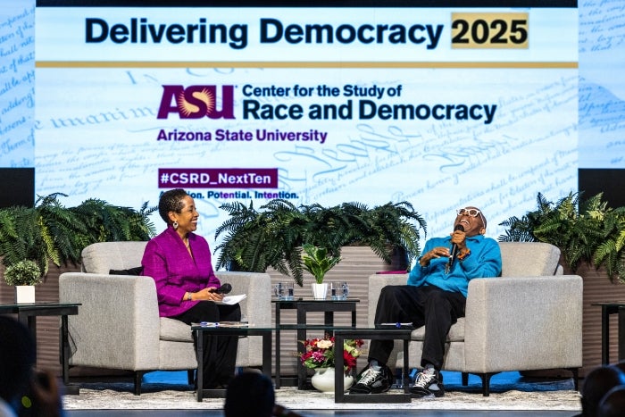 A black woman and man sitting in chairs on a stage with a projection screen behind them