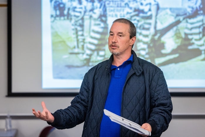 Man with short brown hair wearing a gray jacket and a blue polo shirt speaks in front of a projection screen in a classroom