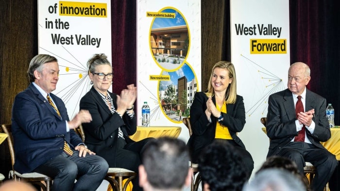 ASU President Crow and Governor Hobbs sit in chairs next to others on stage with West campus posters behind them.