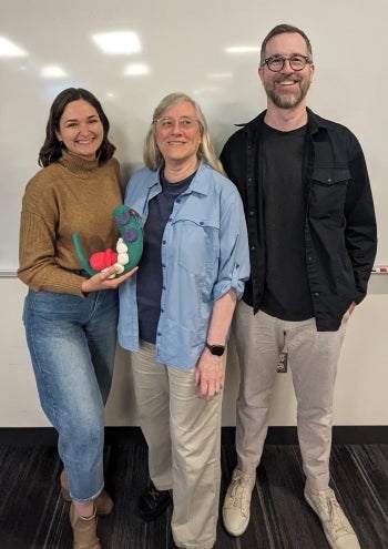 Three smiling people in front of a whiteboard. The woman on the left is holding a clay model of an embryo.