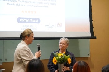 A woman with a microphone stands next to a woman holding flowers.