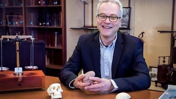 Jonathan Gewirtz smiles and holds a brain model while seated at a table with a balance scale and skull models. Shelves filled with books are in the background.