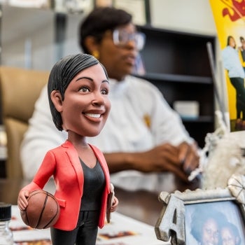 A bobblehead of a black woman with short hear wearing a red blazer and holding a basketball sits on a desk with the woman it represents sitting behind the desk