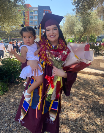 Dani Bermudez in her cap and gown holds her daughter and a bouquet of flowers outdoors.