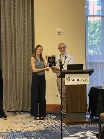 Two people hold a plaque in front of a podium labeled “ADHD 2024.”