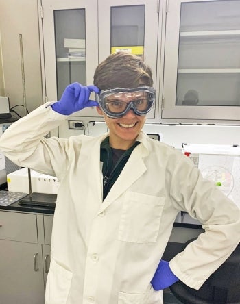 A woman posing in a lab coat wearing blue gloves and safety goggles