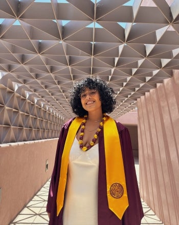 Danielle Ochoa, wearing her ASU graduation robes and a white dress, smiles at the camera while posing under a geometric walkway. 