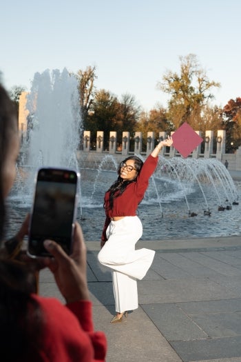 Person taking a photo of a woman posing with her graduation cap in the air in front of a fountain