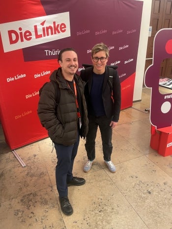 Two people pose for a photo in front of a display with German words on it