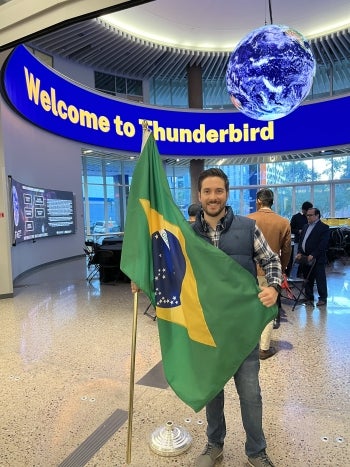 Guilherme holding a Brazilian flag indoors under a sign that reads "Welcome to Thunderbird."