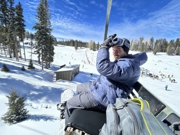 Person on a ski lift, wearing a blue jacket and goggles, with a snowy landscape and ski slopes in the background.