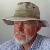 Portrait of a man with a white beard wearing a khaki brimmed hat and a white and blue striped button down shirt