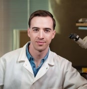 A man in a lab coat sits in front of a microscope, looking at the camera.