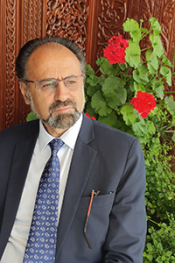 Dr. Mohammad Yousof Asefi sits in front of an elaborately carved wooden door and red flowers wearing a grey suit, blue tie and glasses.